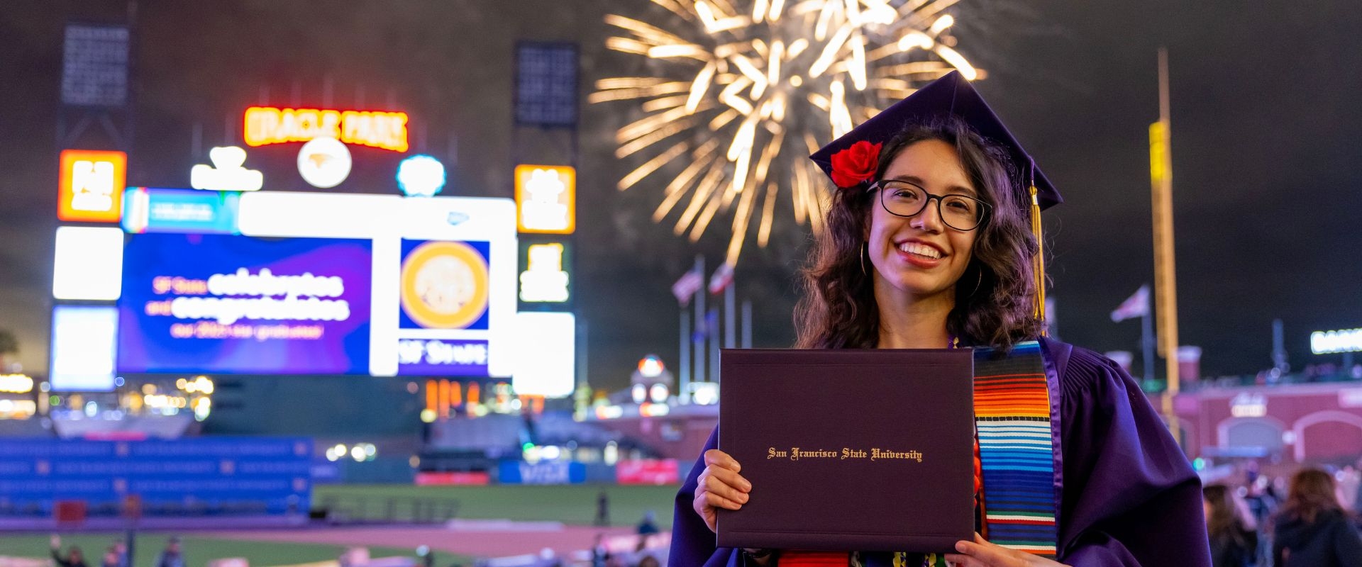 Graduate at Commencement holds up her diploma while fireworks explode at Giants stadium